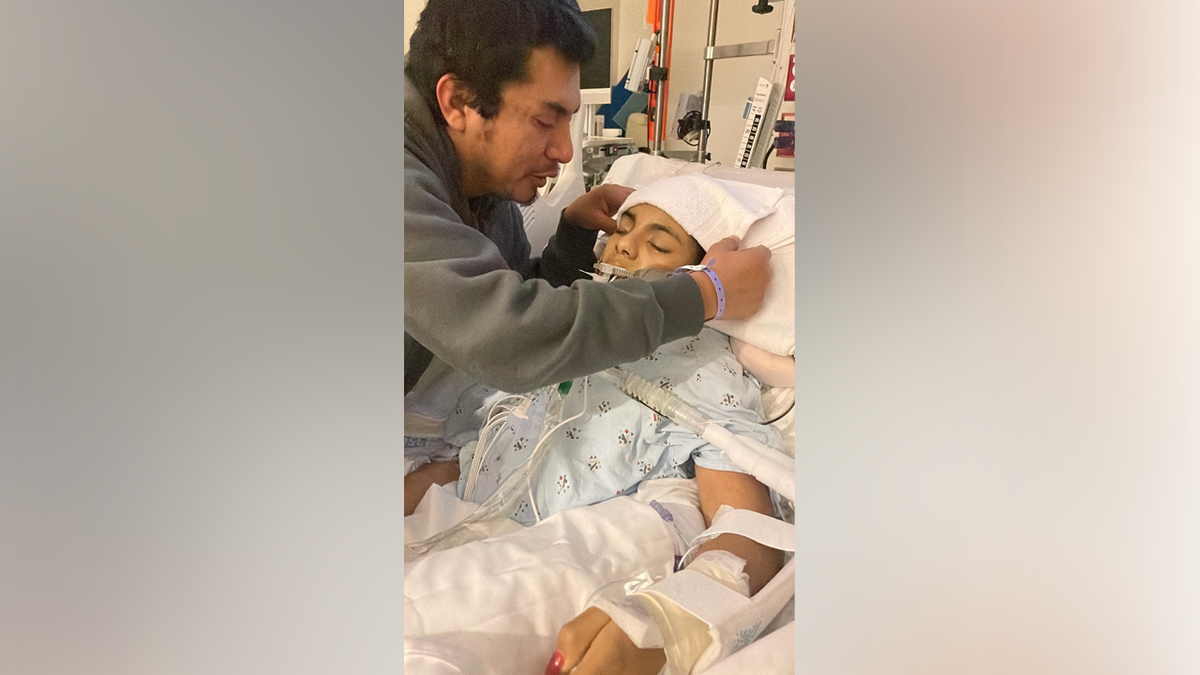 A family member sits beside Khimberly Zavaleta in a hospital bed after she suffered a traumatic brain injury.