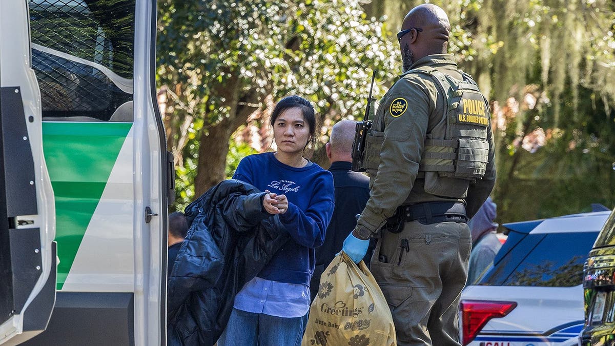 A woman removes her coat next to a US Border Patrol Agent who is holding a bag