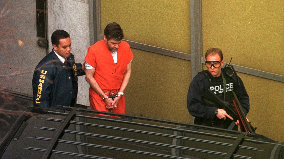 Ted Kaczynski in an orange prison jumpsuit is escorted in handcuffs by law enforcement officers, including one holding a firearm, outside a building.